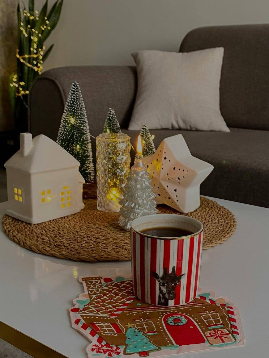 A cozy living room features a white coffee table with a striped mug of coffee, festive gingerbread-themed coaster, mini Christmas trees, and house-shaped candle holders. A sofa with a cushion and fairy lights are in the background.