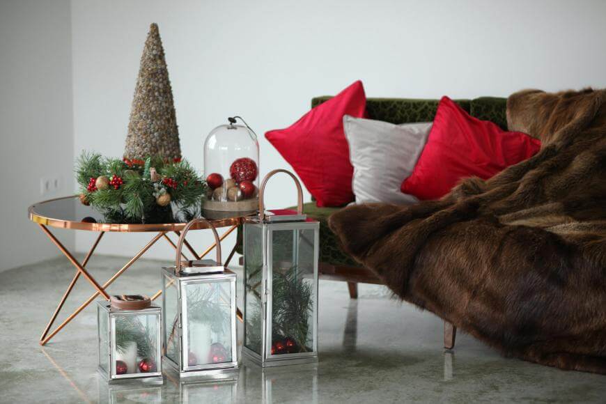 A festive living room features a gold coffee table with Christmas decorations and a faux fur throw on a green sofa with red and silver pillows. Lanterns with ornaments and greenery sit on the polished floor.