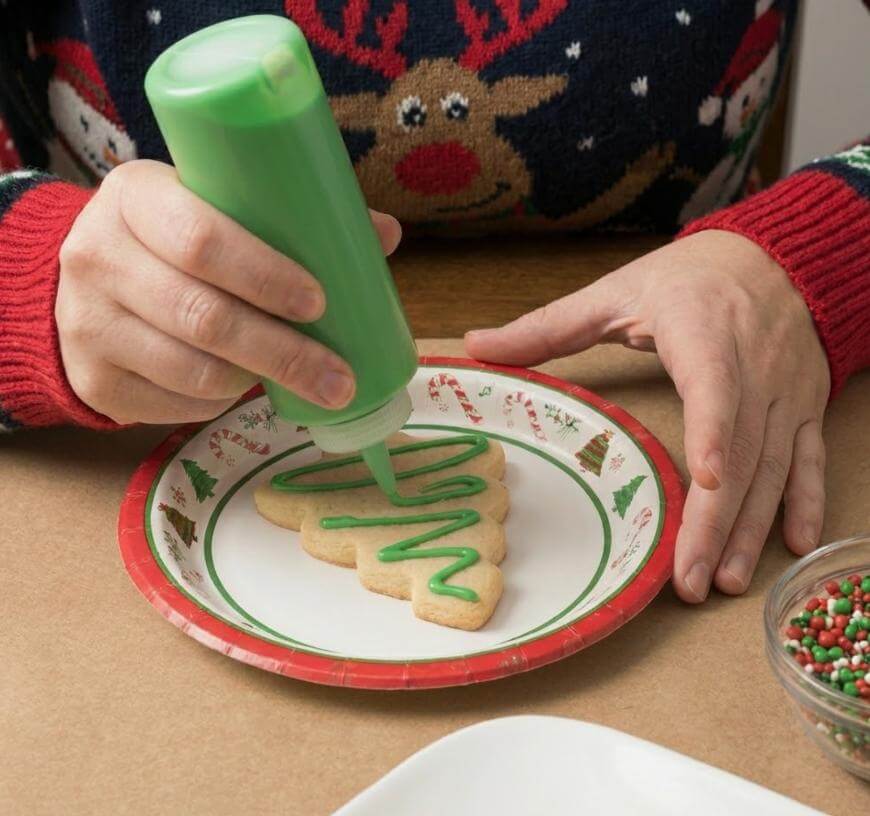 A person in a festive sweater decorates a Christmas tree-shaped cookie with green icing, using a squeeze bottle. The cookie is on a holiday-themed plate, with sprinkles and other cookies nearby.