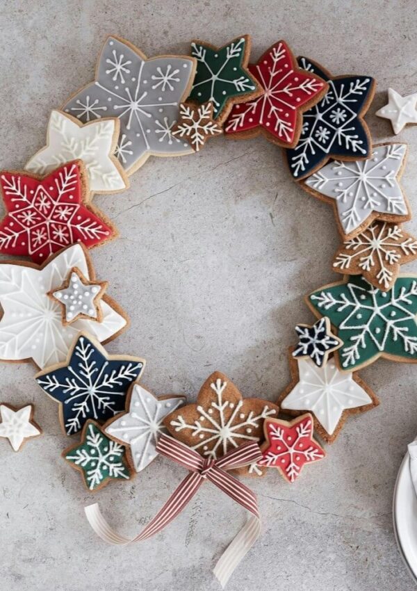 A wreath made of leaf- and star-shaped cookies decorated with colorful icing and white snowflake patterns, arranged in a circle and tied at the bottom with a striped ribbon.