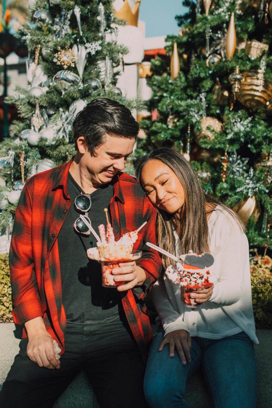 A smiling couple sits together outdoors in front of decorated Christmas trees, holding festive holiday desserts topped with whipped cream, sprinkles, and a Mickey Mouse-shaped cookie.