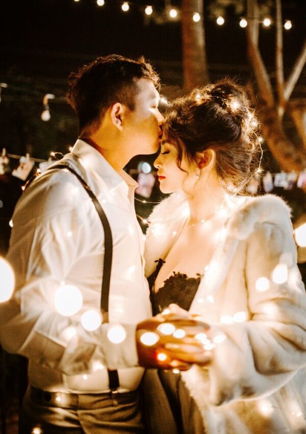 A man in a white shirt kisses a womans forehead as she closes her eyes, both surrounded by warm string lights at night. The woman wears a white fur coat and the scene feels romantic and intimate.