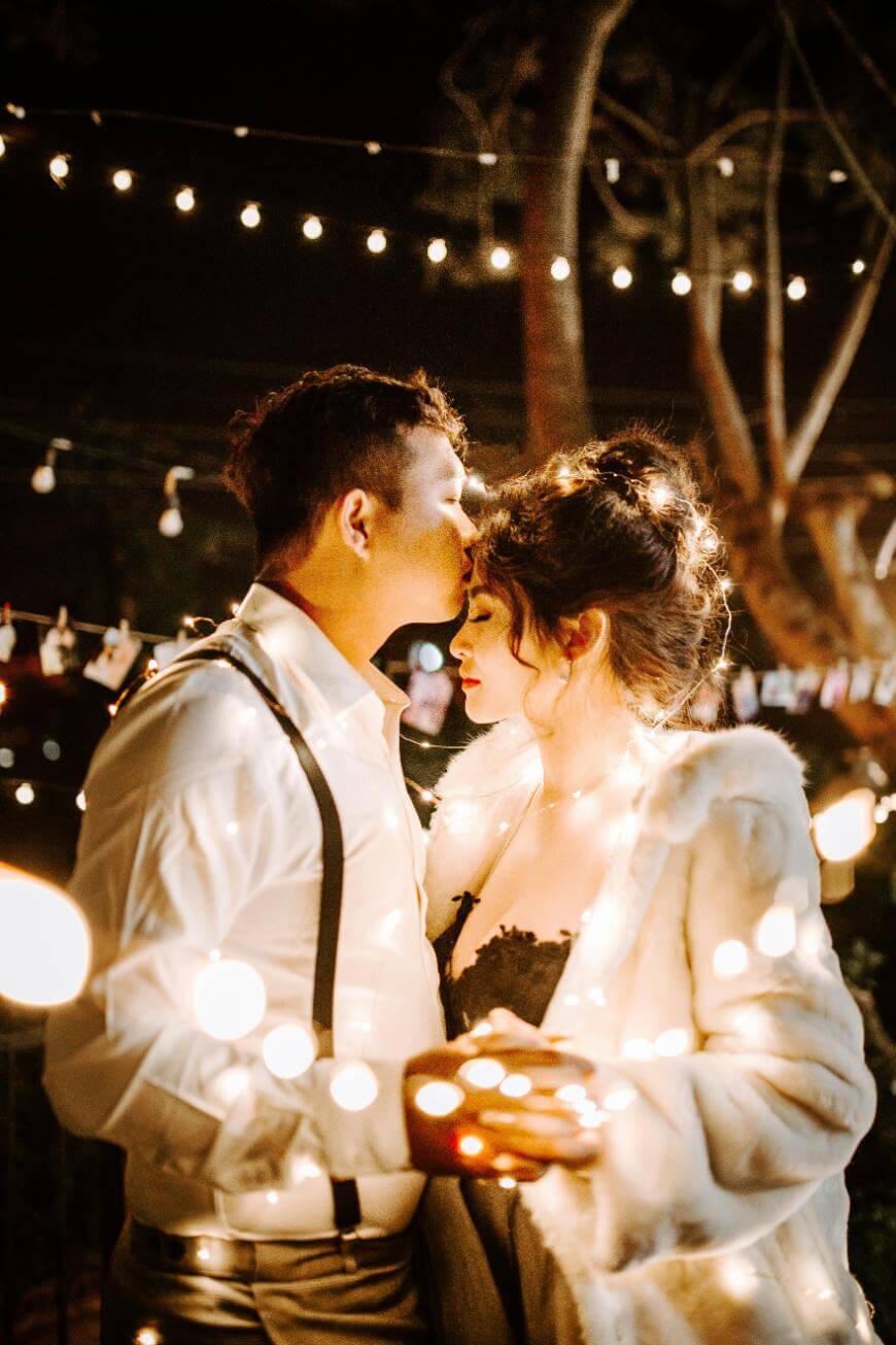 A couple stands close together outdoors at night, surrounded by string lights. The man kisses the woman’s forehead while she holds glowing lights in her hands. They are dressed formally, creating a romantic atmosphere.