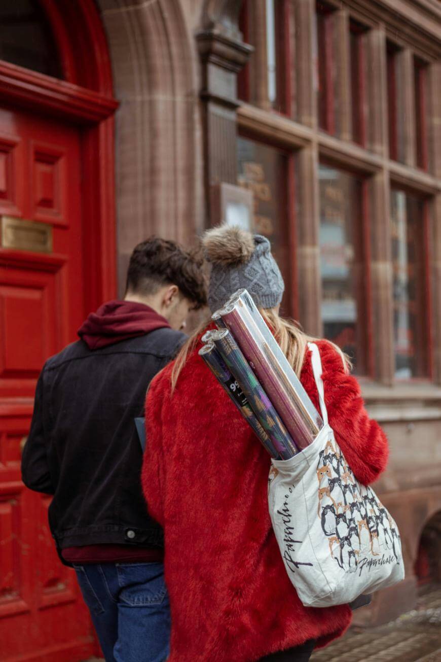 Two people walk next to a building with large windows and a red door. One person wears a red coat and grey hat, carrying rolled posters in a patterned tote bag over their shoulder.