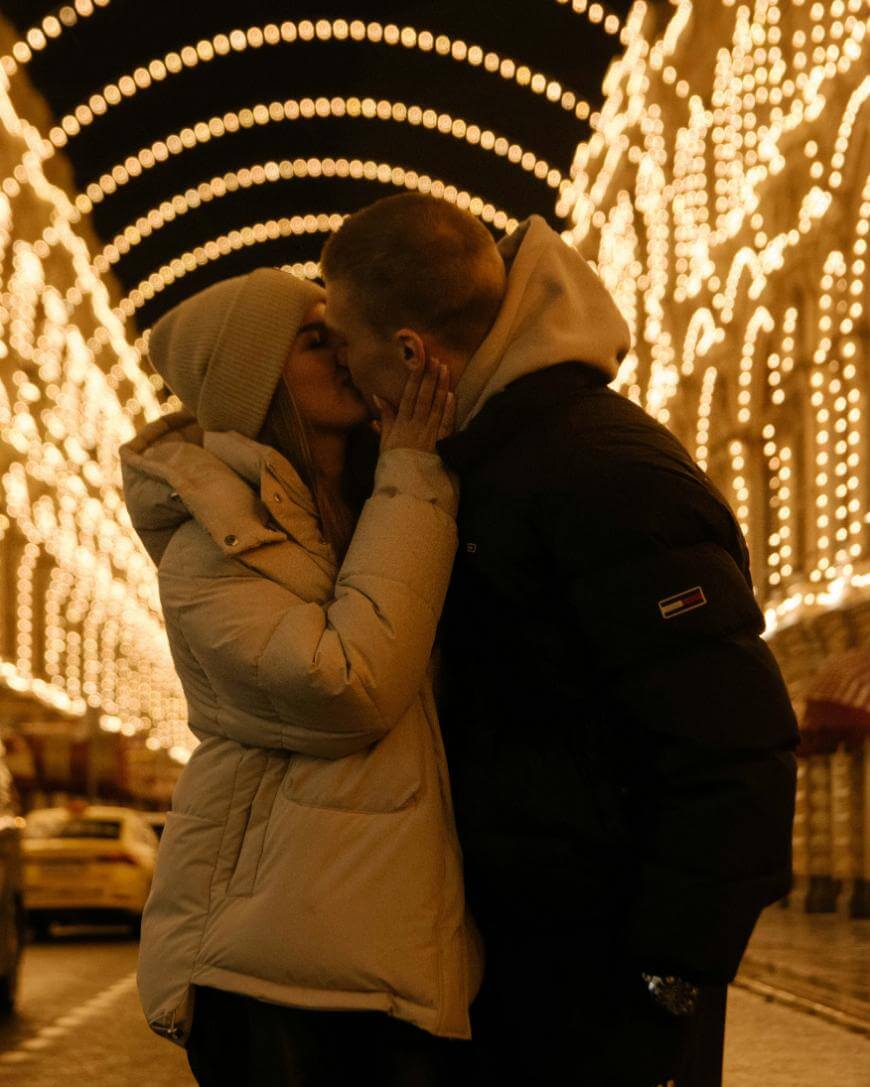 A couple in winter jackets kisses under rows of golden string lights at night, surrounded by illuminated arches on a festive city street.