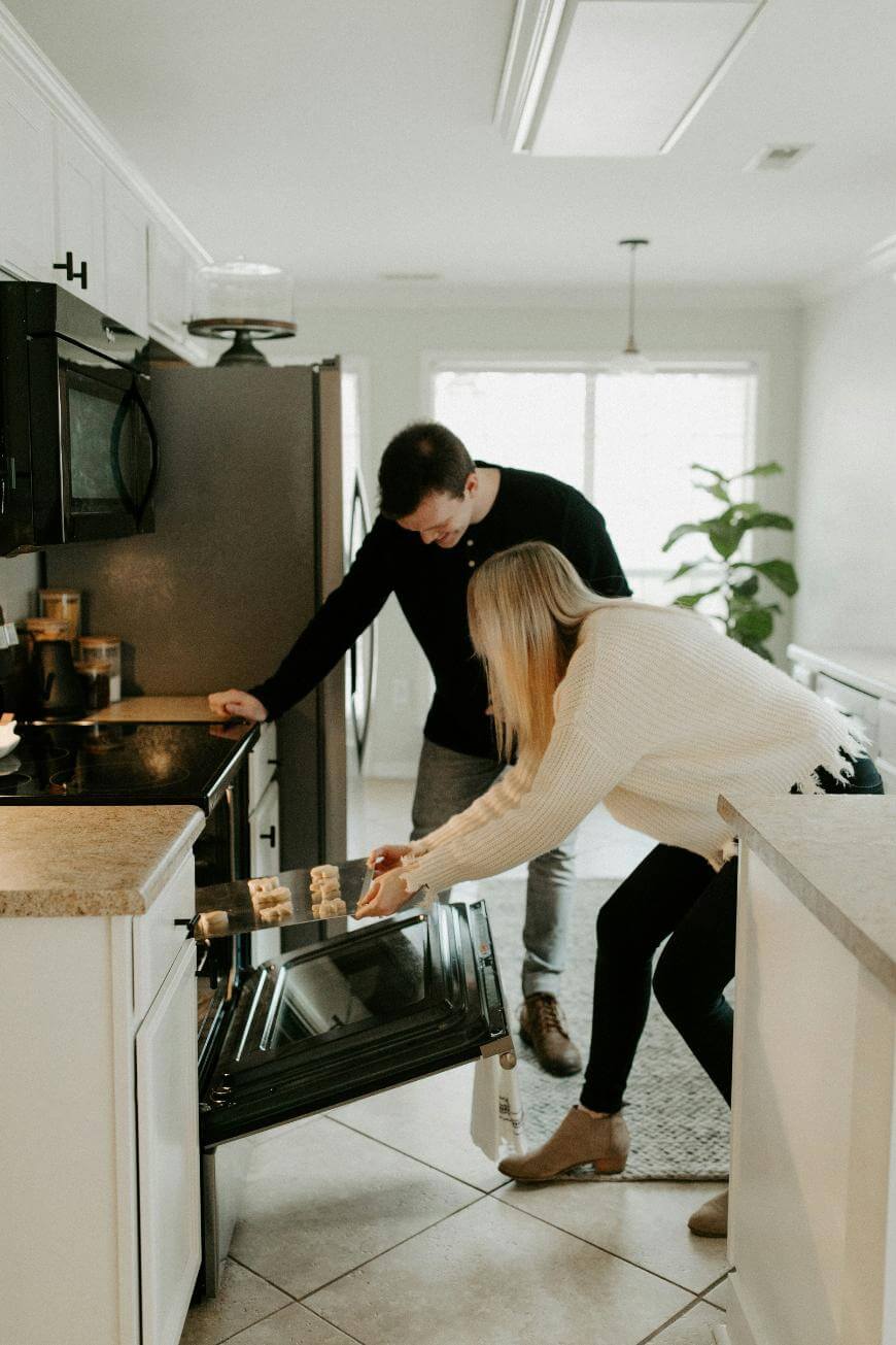 A woman places a tray of food into an open oven while a man stands beside her in a bright, modern kitchen.