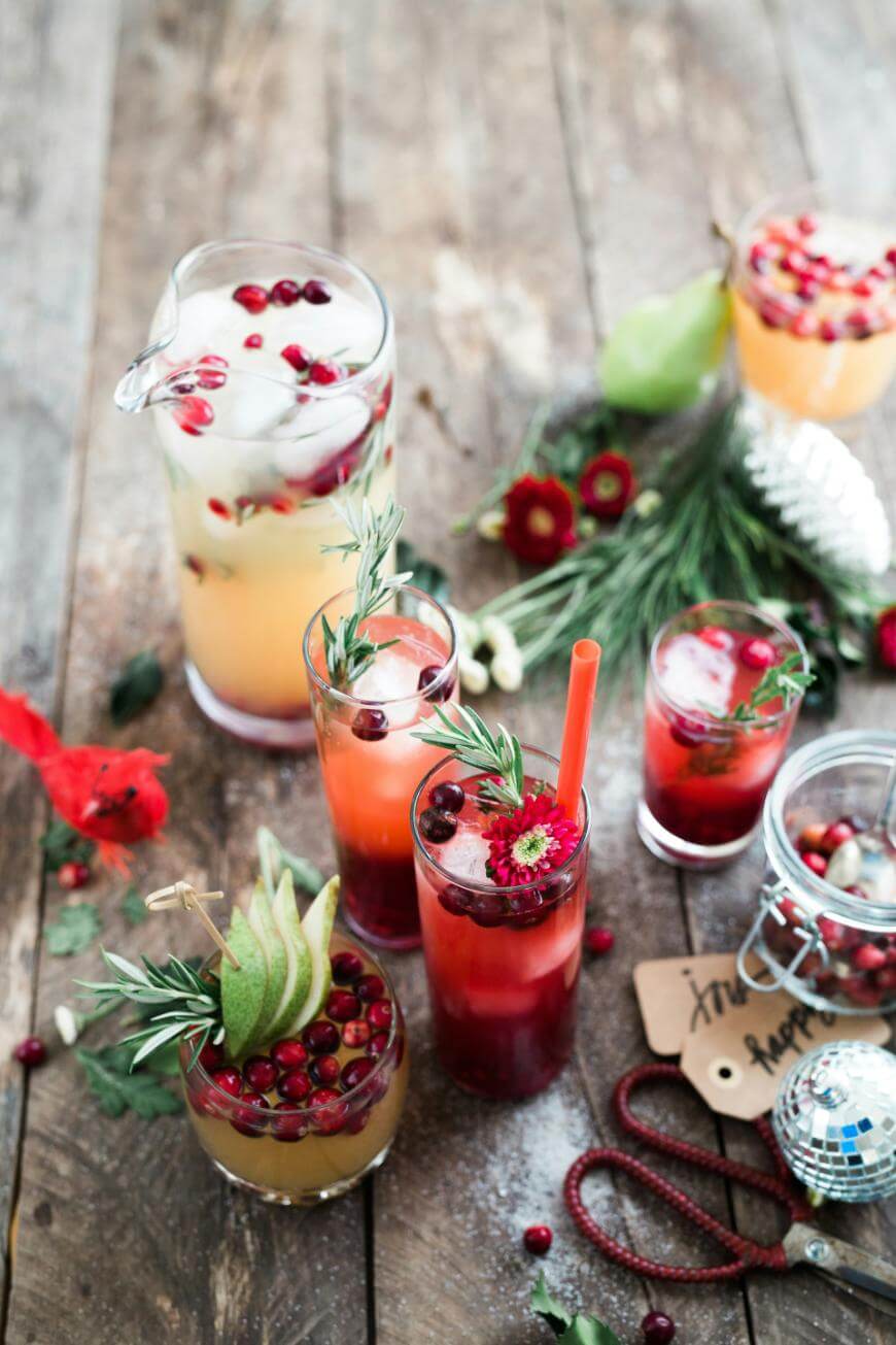 A rustic wooden table with festive drinks in glasses and a pitcher, garnished with cranberries, pear slices, rosemary, and flowers. Holiday decorations and greenery are scattered around.