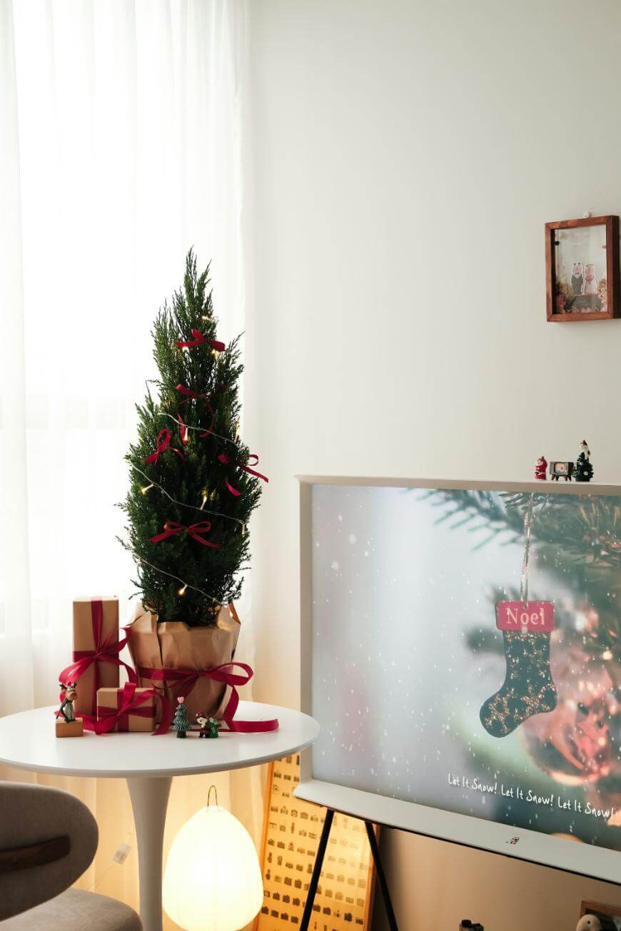 A small Christmas tree with red ribbons and gifts on a round white table beside a TV displaying a festive Noel stocking. A warm lamp glows below, and a framed photo hangs on the wall.