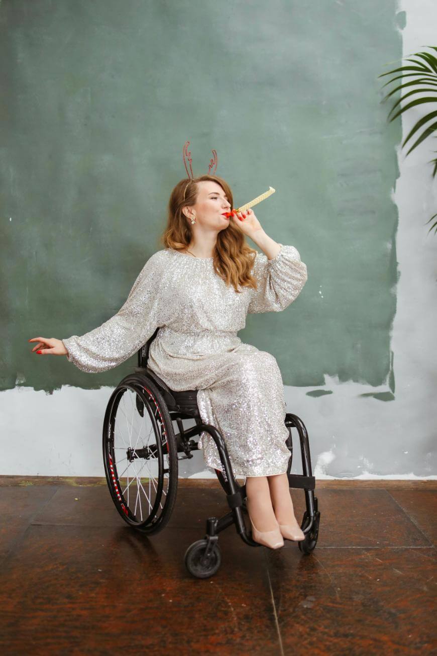 A woman in a sparkling silver dress sits in a wheelchair, wearing reindeer antlers and blowing a party horn. She appears joyful, with a festive and celebratory expression, in front of a plain green backdrop.
