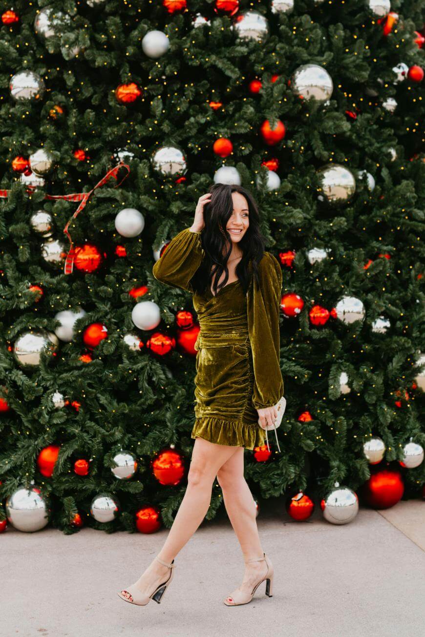 A woman in a green velvet dress and nude heels poses and smiles in front of a large, decorated Christmas tree with red and silver ornaments.