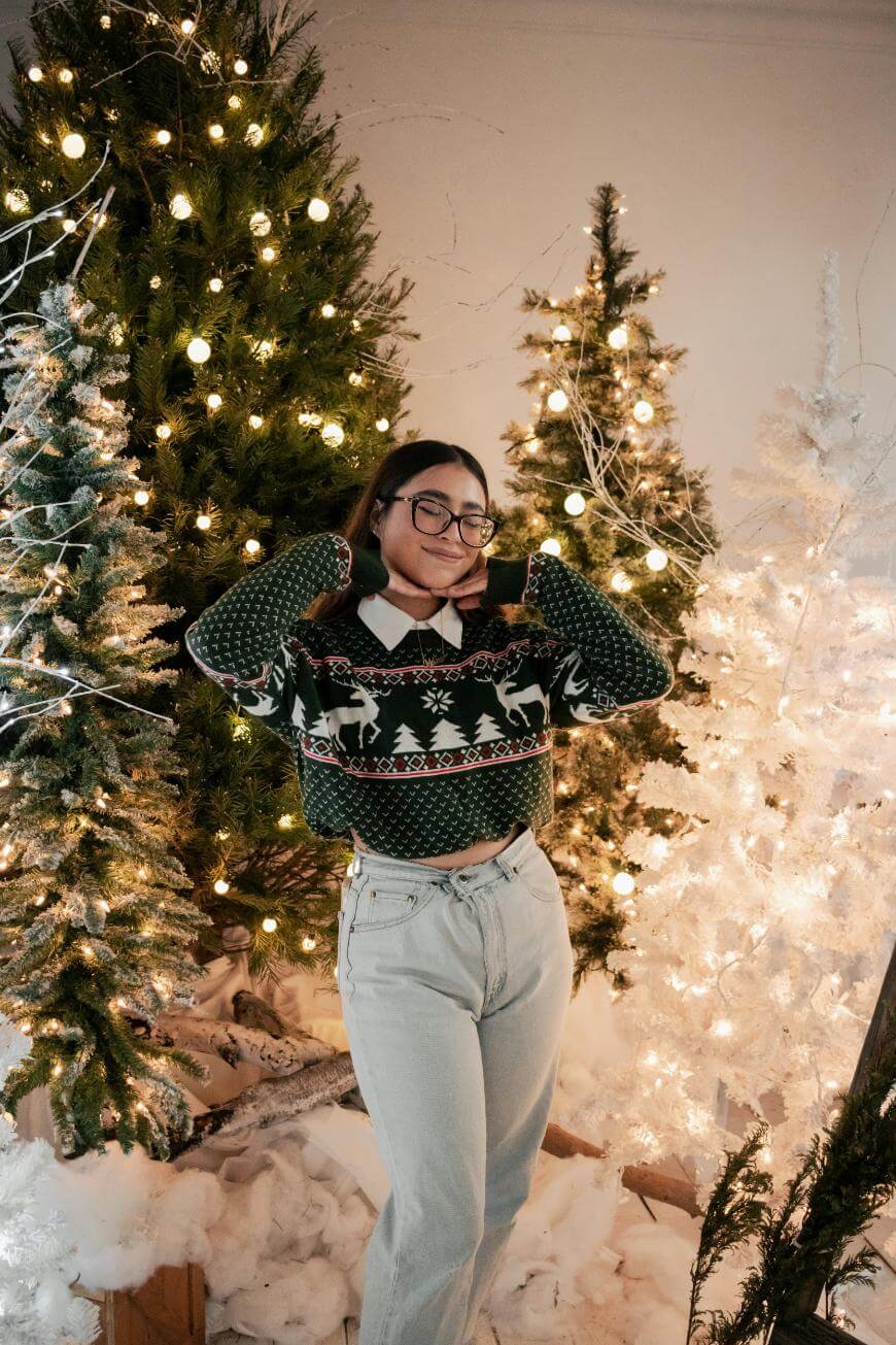 A person wearing glasses, a festive sweater with reindeer, and light jeans poses smiling in front of lit Christmas trees surrounded by fake snow and holiday decorations.