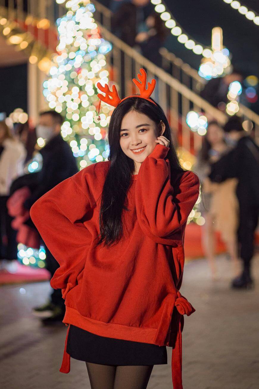 A young woman in a red sweater and reindeer antler headband smiles and poses in front of a lit-up Christmas tree, with festive lights and people in the background.