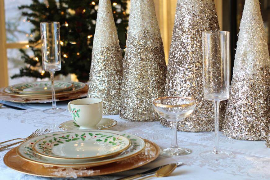 A festive table setting with gold-rimmed plates, glassware, and a tea cup on a white tablecloth. Glittery cone decorations and a Christmas tree with lights are visible in the background.