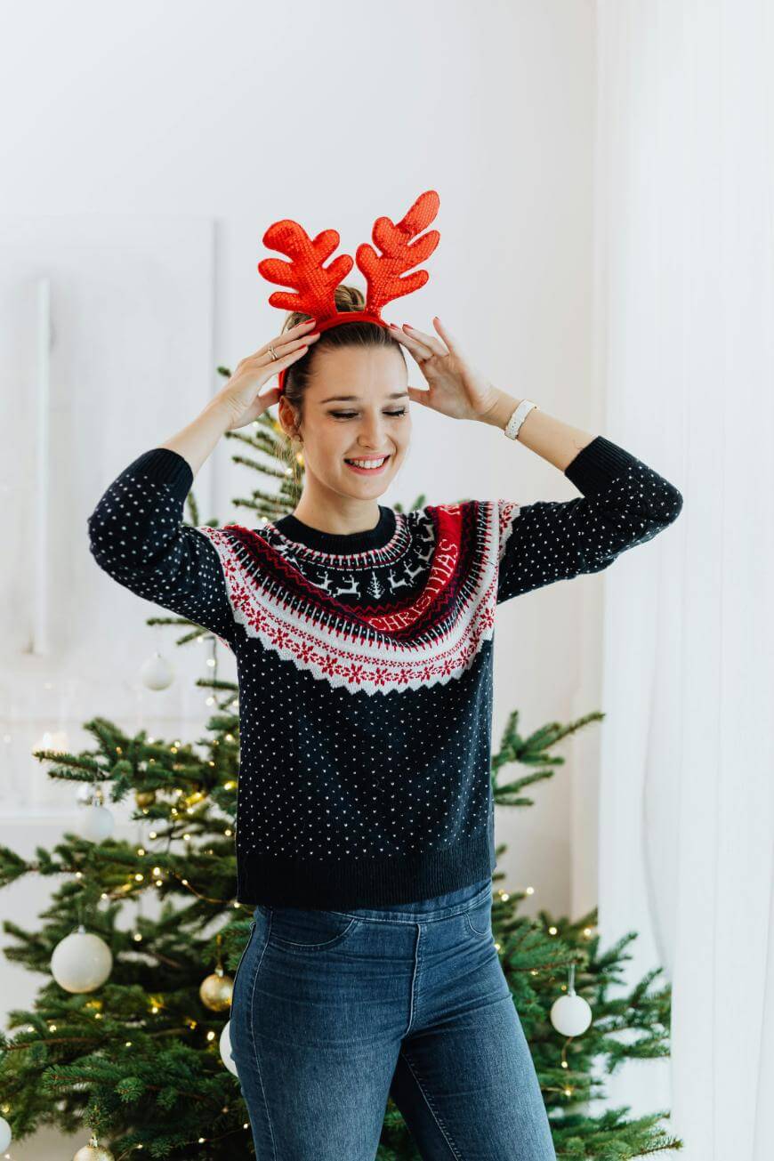 A smiling woman wearing a festive sweater and red reindeer antlers stands in front of a decorated Christmas tree with white ornaments and lights.