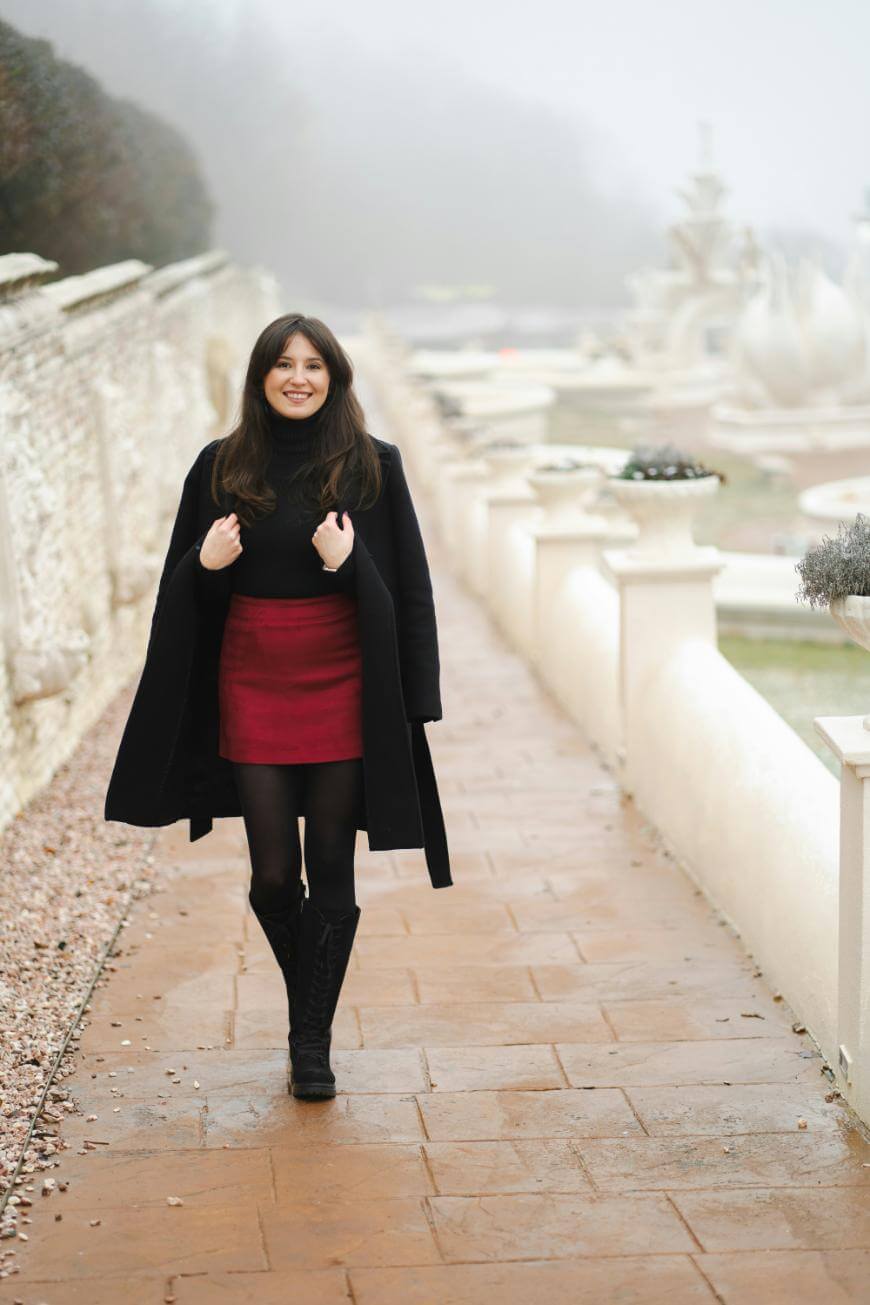 A woman wearing a black coat, black top, red skirt, black tights, and boots walks along a paved garden path bordered by a stone wall and white decorative planters on a foggy day.