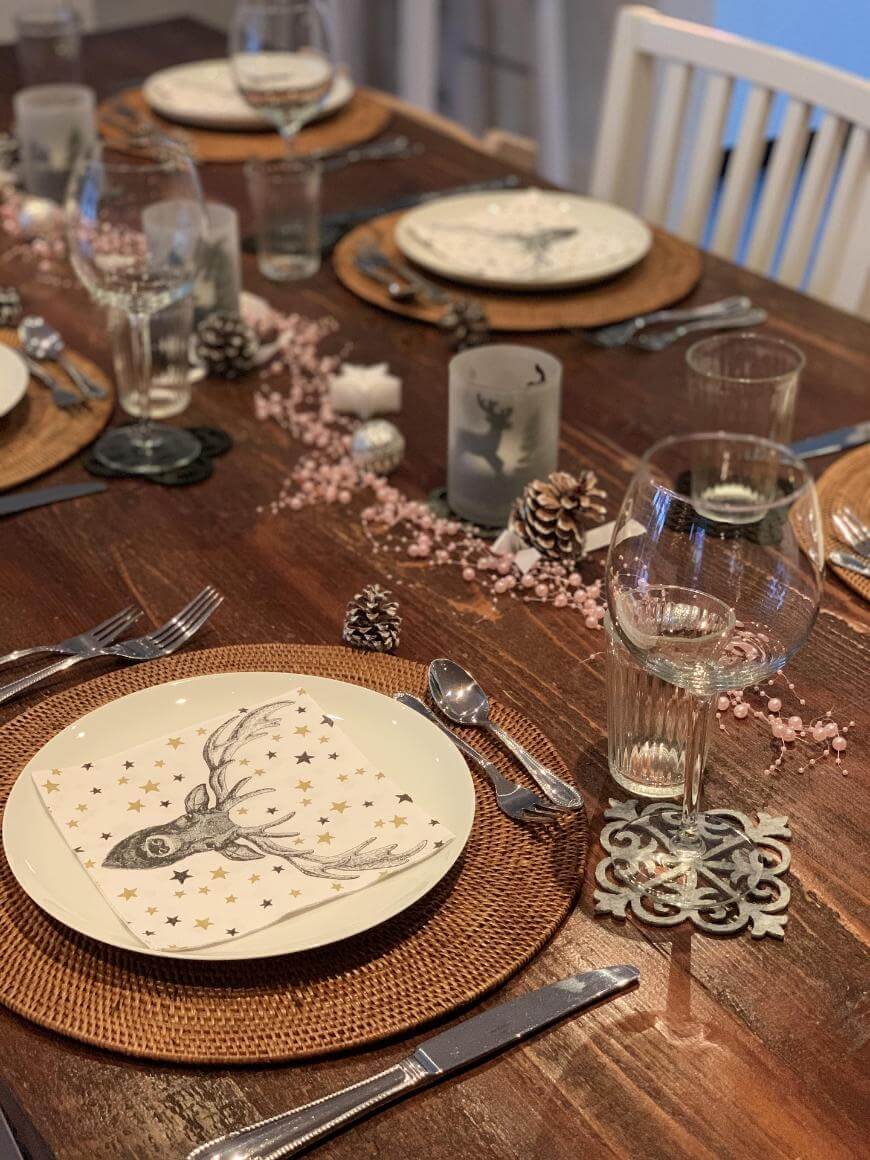 A wooden dining table set for a meal with woven placemats, deer-themed plates, wine glasses, cutlery, and festive decorations including pinecones and pink beads, creating a cozy, rustic atmosphere.