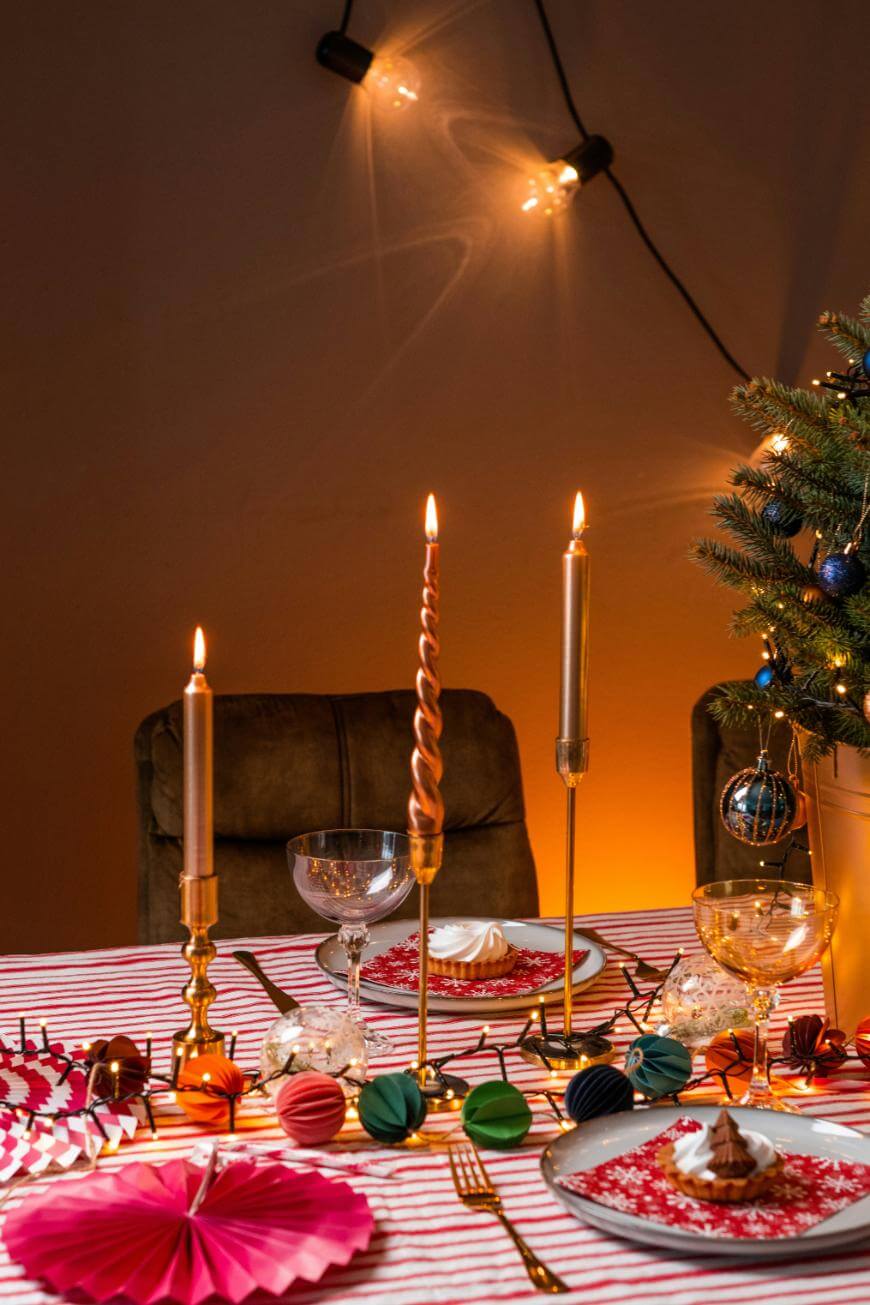 A festive table setting with lit candles, holiday decorations, and a small Christmas tree. The table has a red-and-white striped cloth, goblets, plates with desserts, and colorful paper ornaments, with warm string lights in the background.
