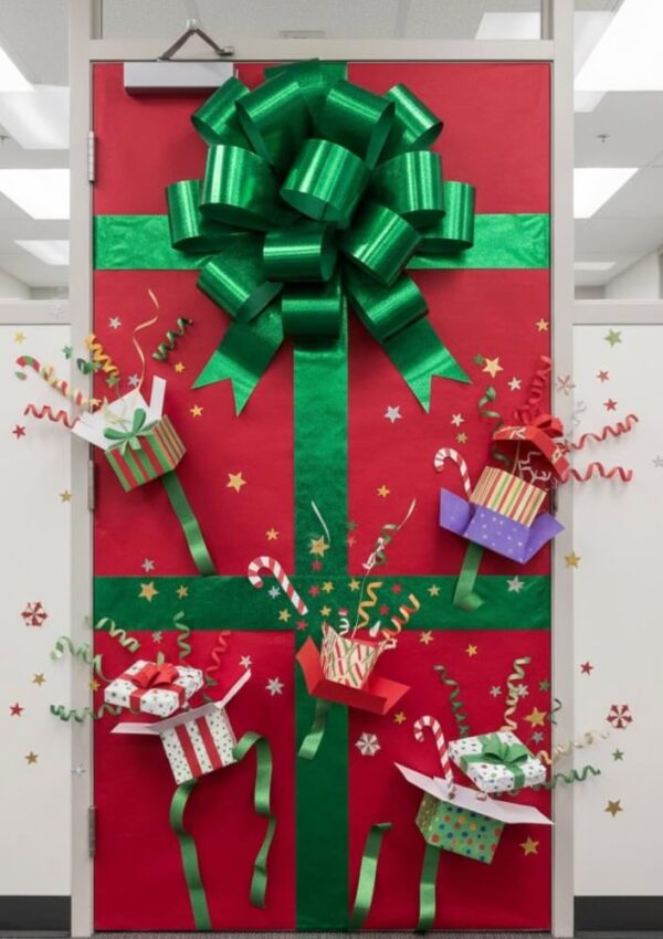 A door decorated like a large gift with red paper and green ribbon, topped with a big green bow. Colorful pop-out presents and shiny stars are attached, creating a festive holiday display.