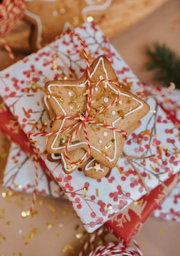 Three festively wrapped gifts are stacked, topped with tied star-shaped cookies decorated with white icing, surrounded by gold confetti and a sprig of greenery in the background.
