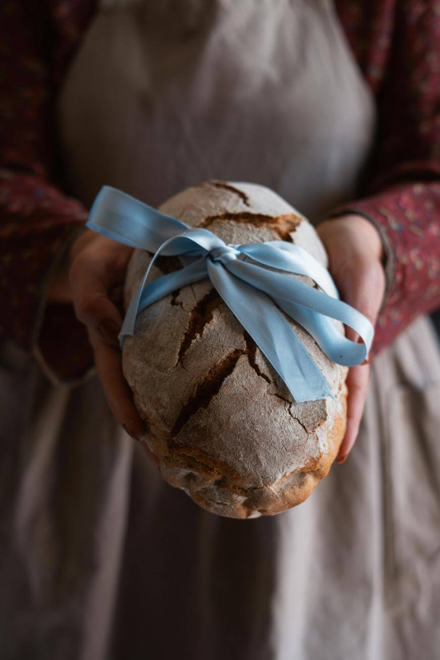 A person wearing a brown apron holds a rustic loaf of bread wrapped with a light blue ribbon, presenting it forward with both hands.