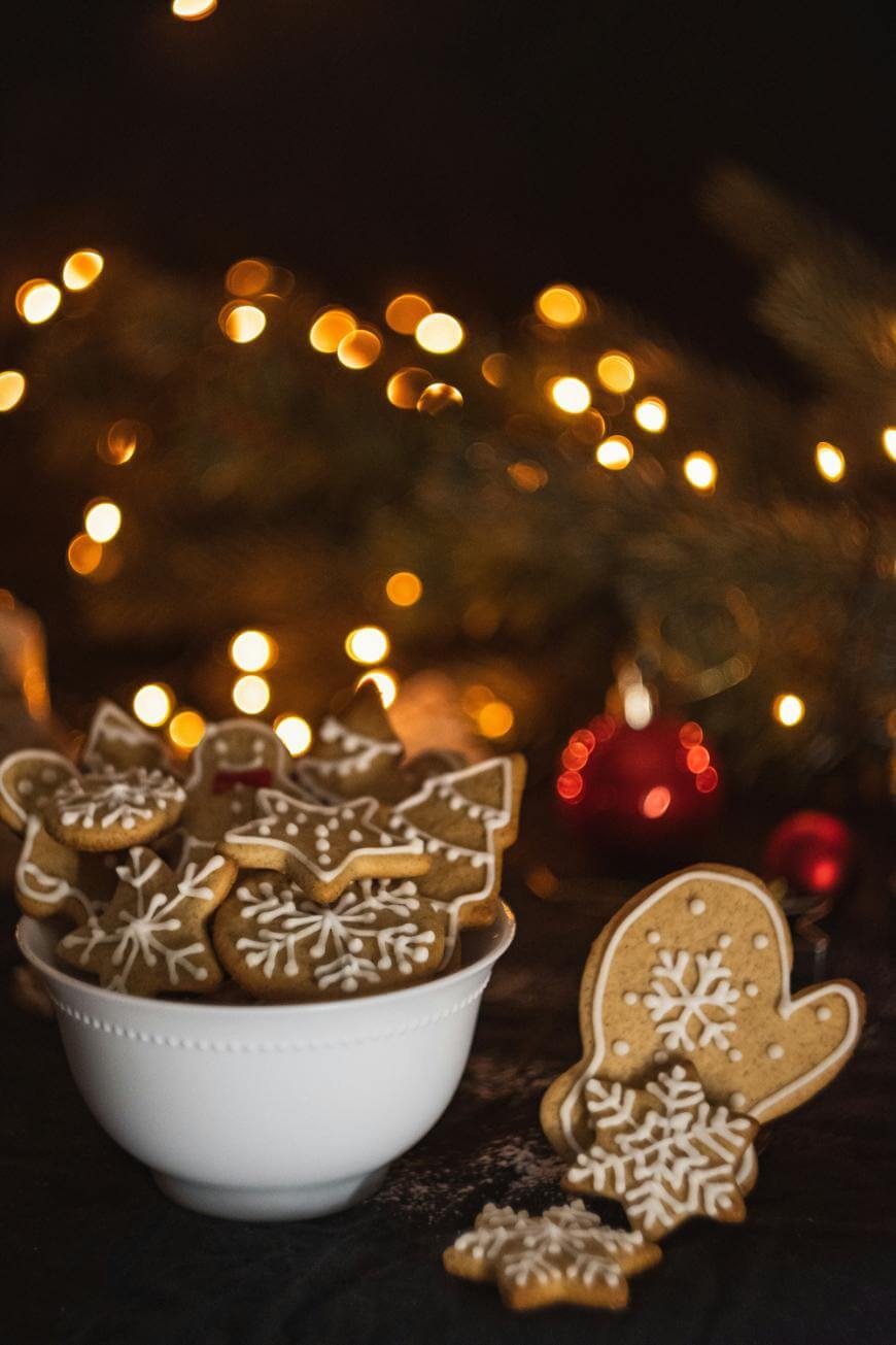 A white bowl filled with decorated gingerbread cookies sits on a table. One mitten-shaped cookie leans against the bowl. Warm, blurred holiday lights and a Christmas tree are visible in the background.