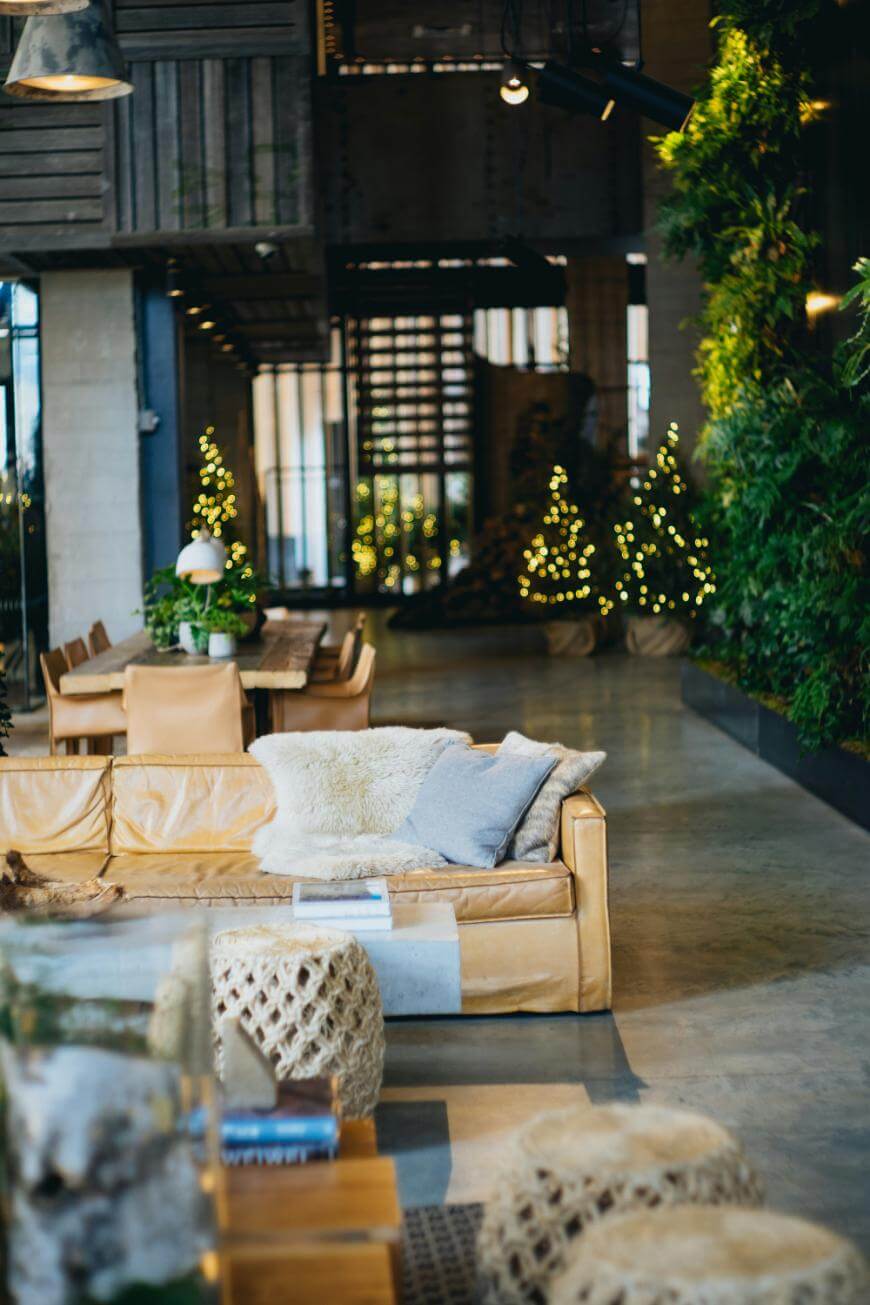 A cozy, modern living space with tan leather sofas, textured cushions, knitted poufs, a long wooden dining table, indoor plants, and small string-lit trees, featuring large windows and an industrial staircase.