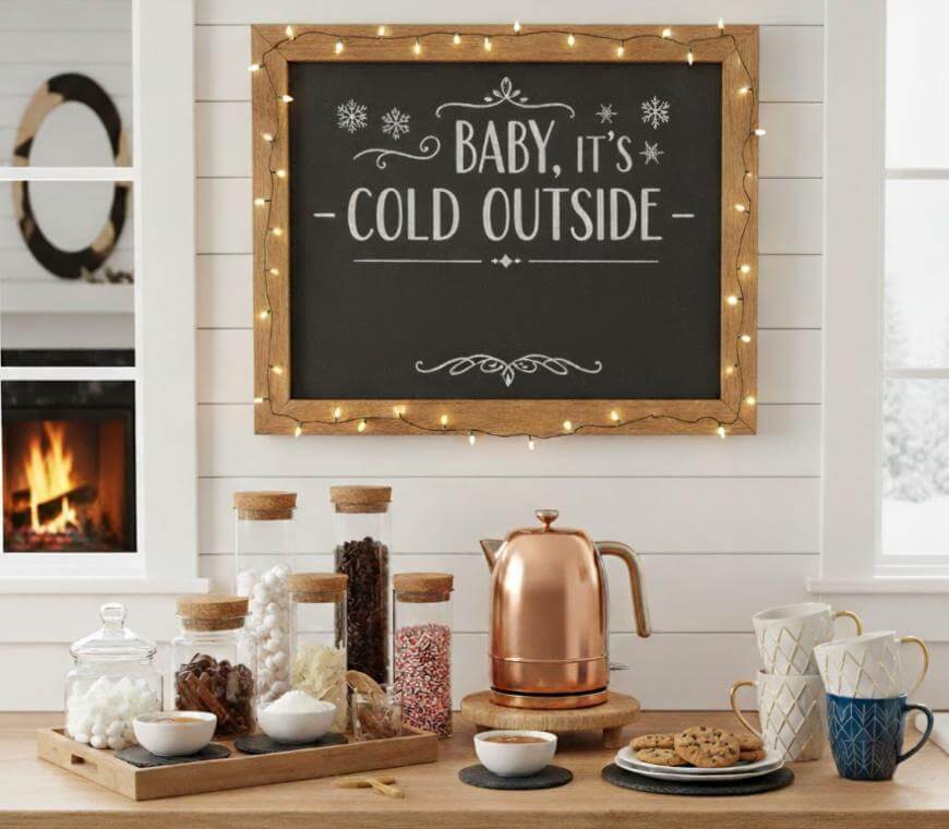 A cozy kitchen scene with a hot drink station, featuring mugs, toppings, a copper kettle, and jars of treats. Above, a chalkboard sign with lights reads Baby, its cold outside. A fireplace glows in the background.