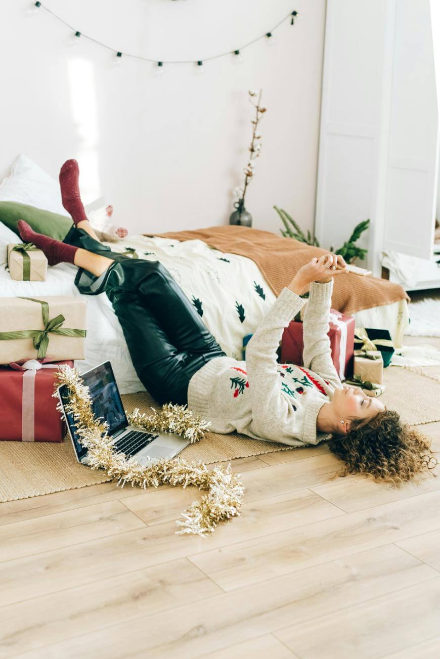 A person wearing festive clothes lies on their back on the floor by a bed, surrounded by wrapped gifts, a laptop, and gold tinsel, appearing relaxed and cheerful in a cozy, decorated room.