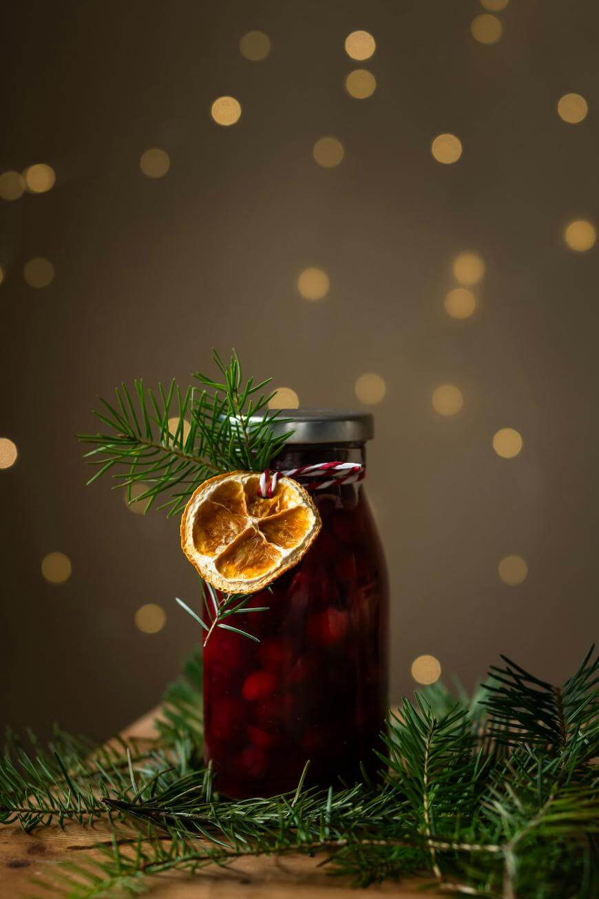 A jar of preserved fruit with a black lid, decorated with a dried orange slice and pine sprig, sits on a wooden surface surrounded by pine branches. Soft golden lights create a festive, blurred background.