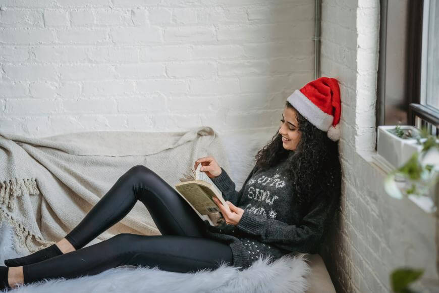 A woman wearing a Santa hat and festive sweater is lounging on a cozy couch by a window, smiling as she reads a book.