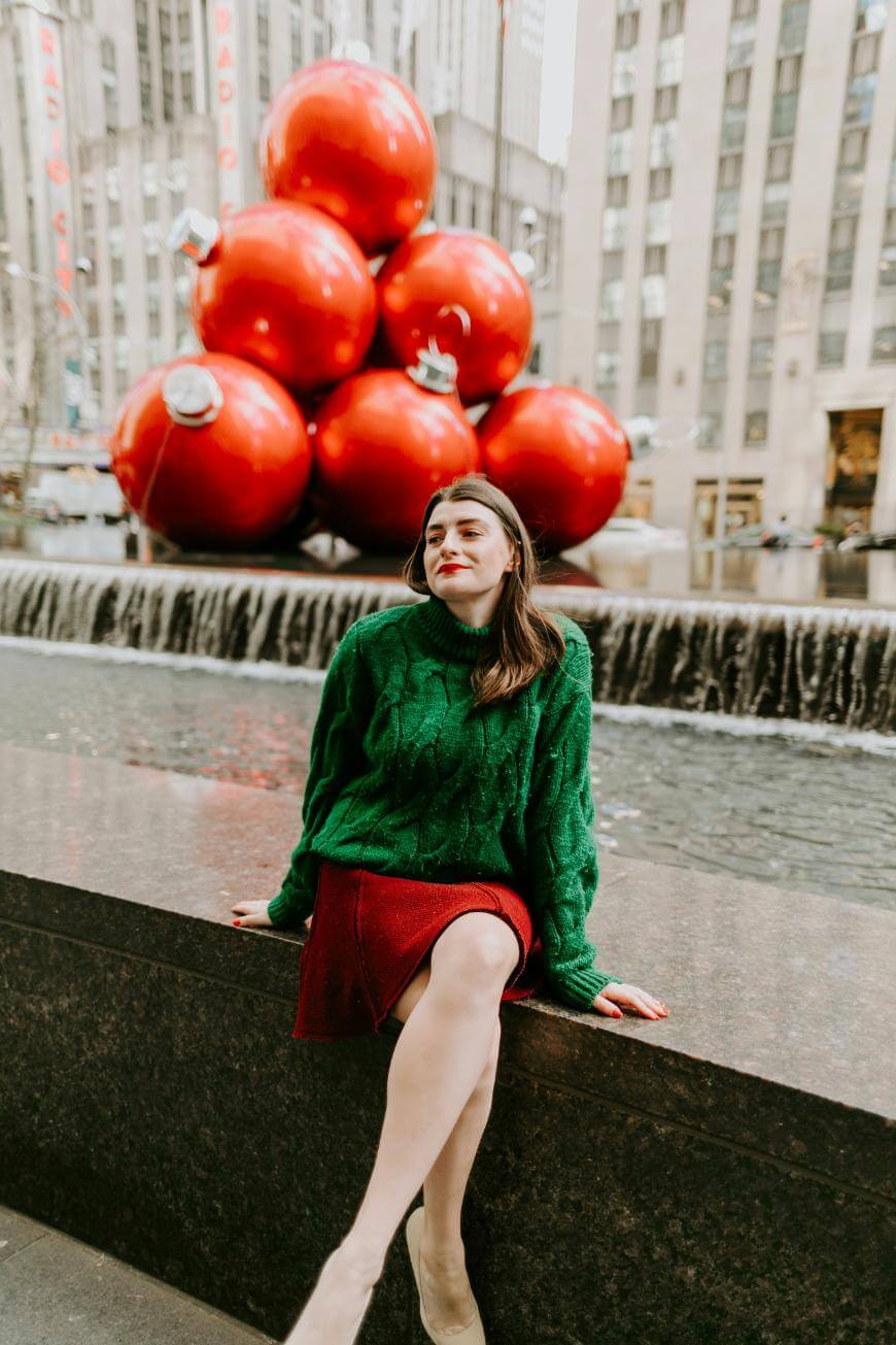 A woman in a green sweater and red skirt sits by a fountain, with large red Christmas ornaments stacked behind her in an urban plaza, likely during the holiday season.