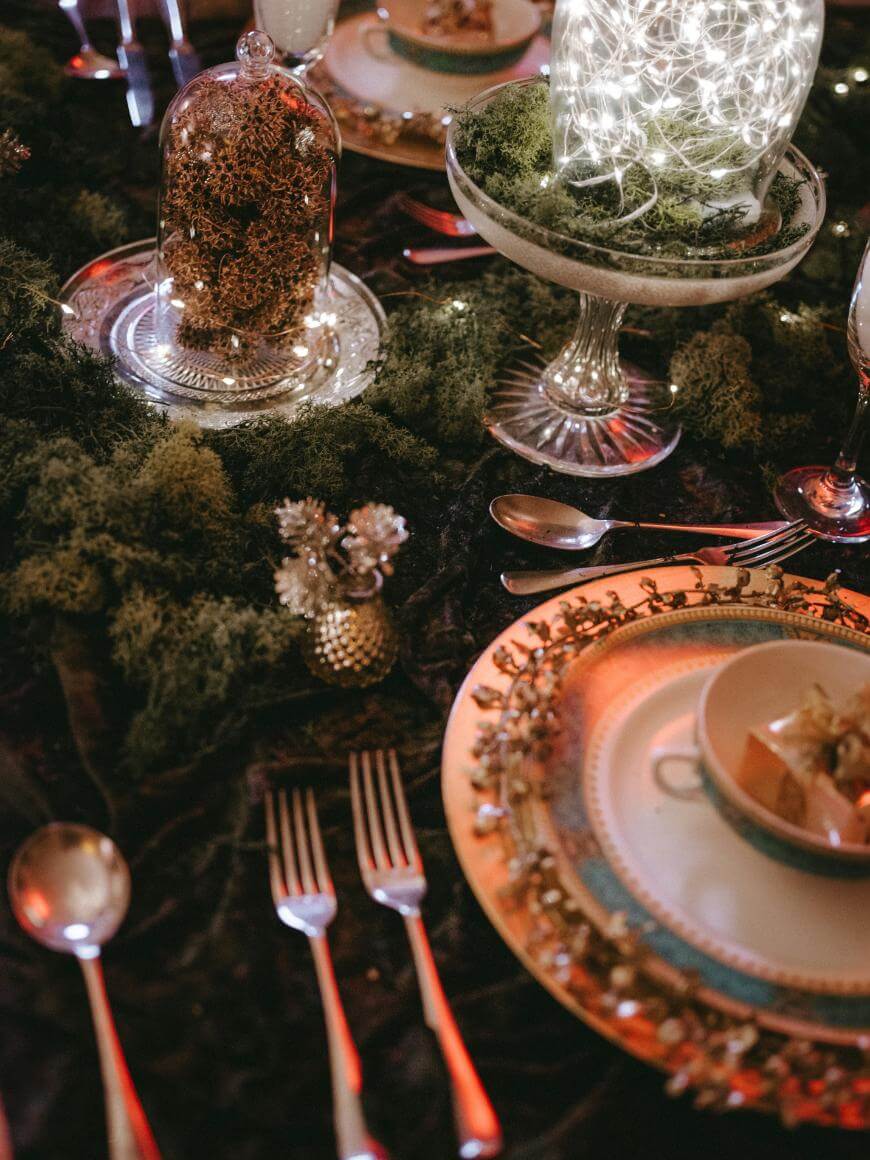 Elegant table setting with ornate plates, bowls, and cutlery on a dark tablecloth, decorated with moss, fairy lights in a glass bowl, and cloches covering pinecones for a festive, nature-inspired look.