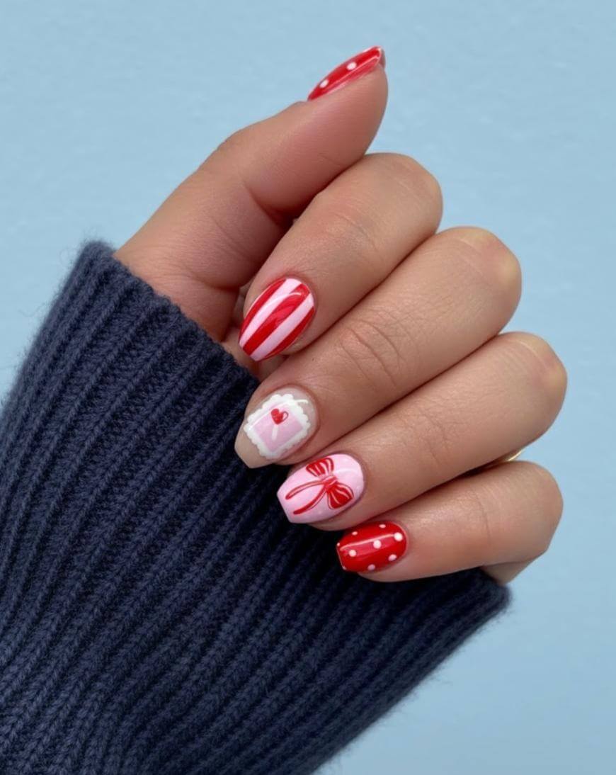 A hand with red, white, and pink themed nail art, featuring stripes, polka dots, a heart, and a bow design. The person is wearing a dark ribbed sweater and is posing against a light blue background.