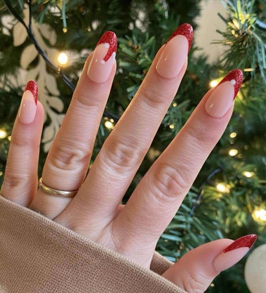 A hand with long almond-shaped nails featuring nude polish and red glittery French tips, held up in front of a lit Christmas tree. The person is wearing a simple gold ring and a beige sleeve.