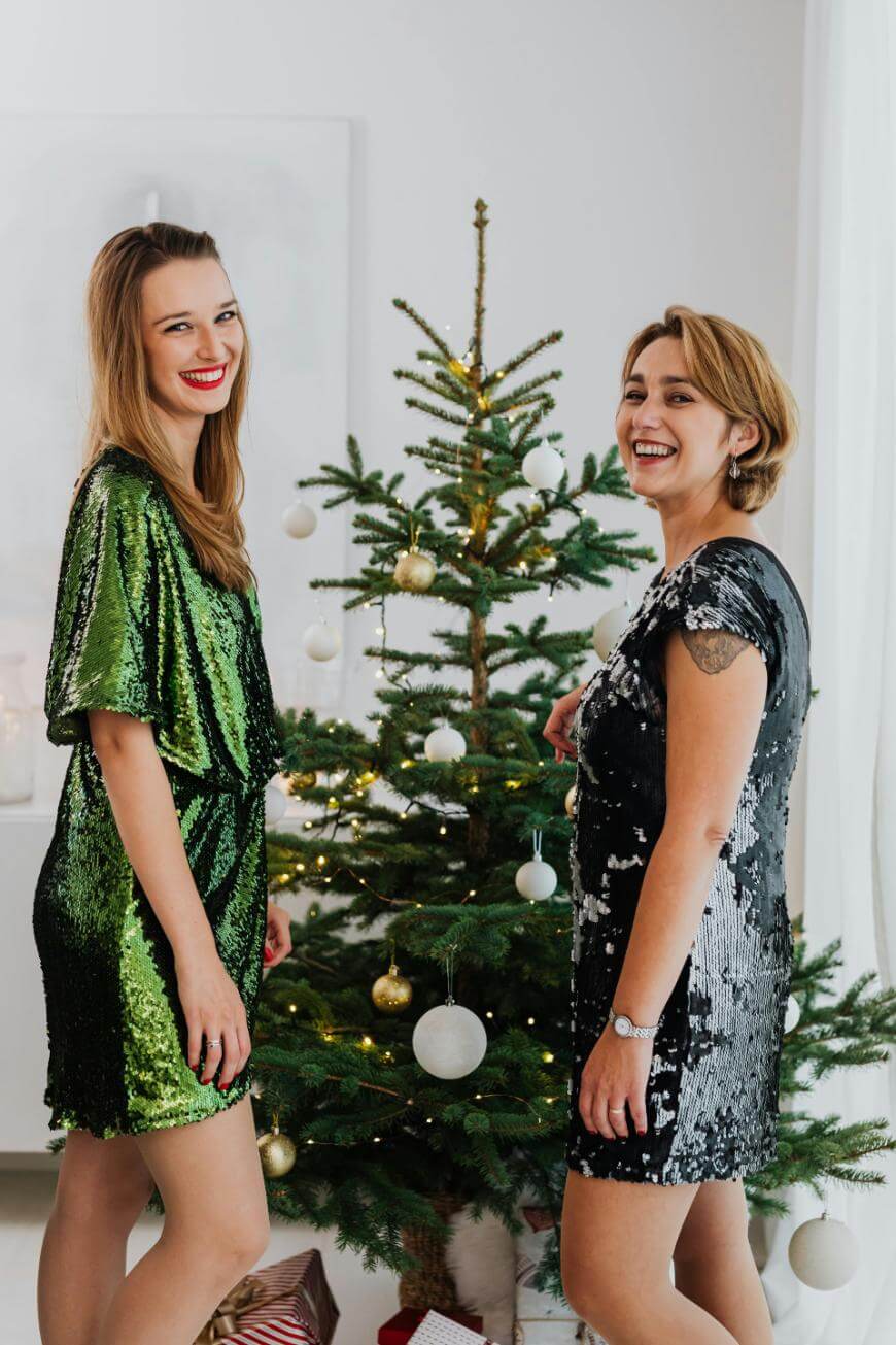 Two women in sparkly dresses smile and stand beside a decorated Christmas tree with lights and ornaments. Wrapped gifts are visible under the tree in a bright, festive room.