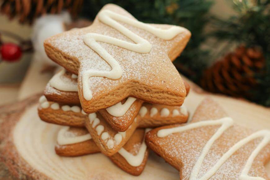 A stack of star-shaped cookies decorated with white icing and powdered sugar sits on a wooden surface, with pinecones and greenery blurred in the background.