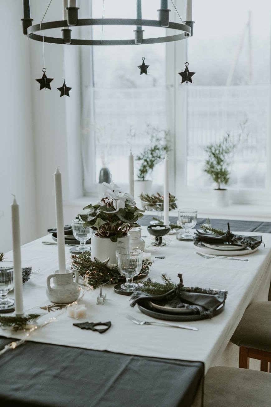 A dining table set for a meal with black and white decor, white candles, glassware, black napkins, and greenery. A chandelier with hanging star ornaments is above, and potted plants are on the windowsill in the background.