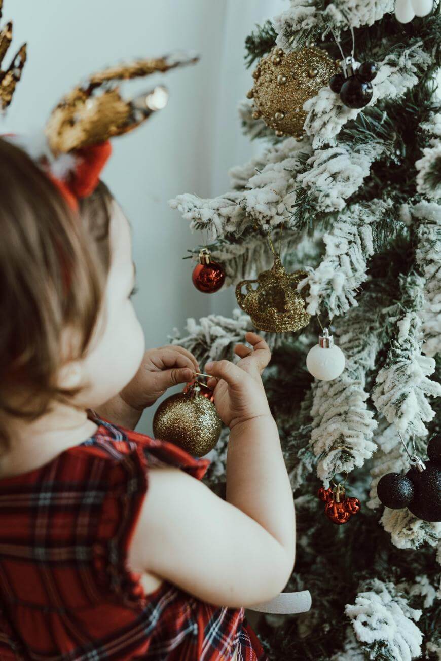 A young child wearing reindeer antlers and a red plaid dress hangs a gold ornament on a snow-covered Christmas tree decorated with baubles.