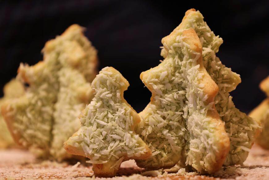 Christmas tree-shaped cookies topped with green-tinted shredded coconut, standing upright on a wooden surface with a dark background.