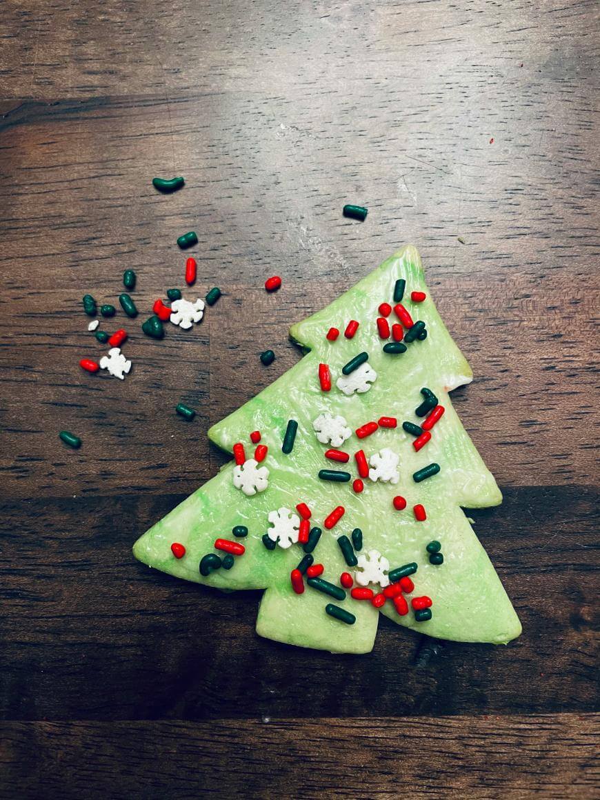Green Christmas tree-shaped cookie topped with red, white, and green sprinkles sits on a dark wooden surface, with extra sprinkles scattered nearby.