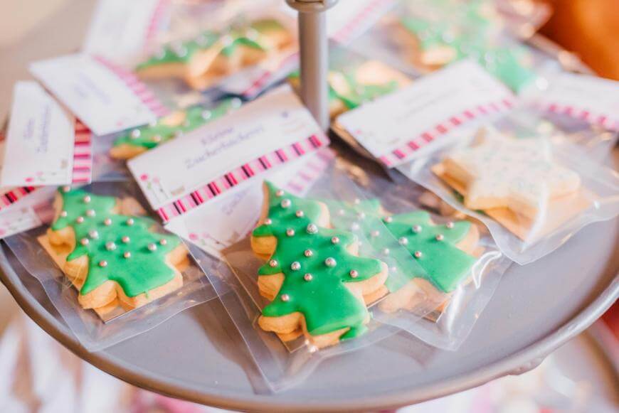 Christmas tree-shaped cookies decorated with green icing and silver sprinkles are individually wrapped in clear plastic and displayed on a round tray. Some star-shaped cookies with white icing are also visible in the background.