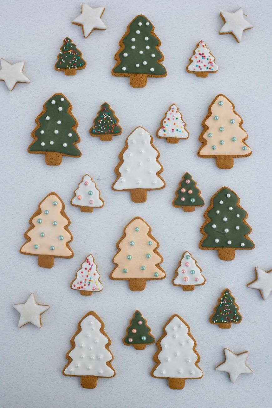 Assorted Christmas tree and star-shaped cookies decorated with white, green, and pastel icing, arranged neatly on a light background. Some cookies have colorful sprinkles or dots as decorations.