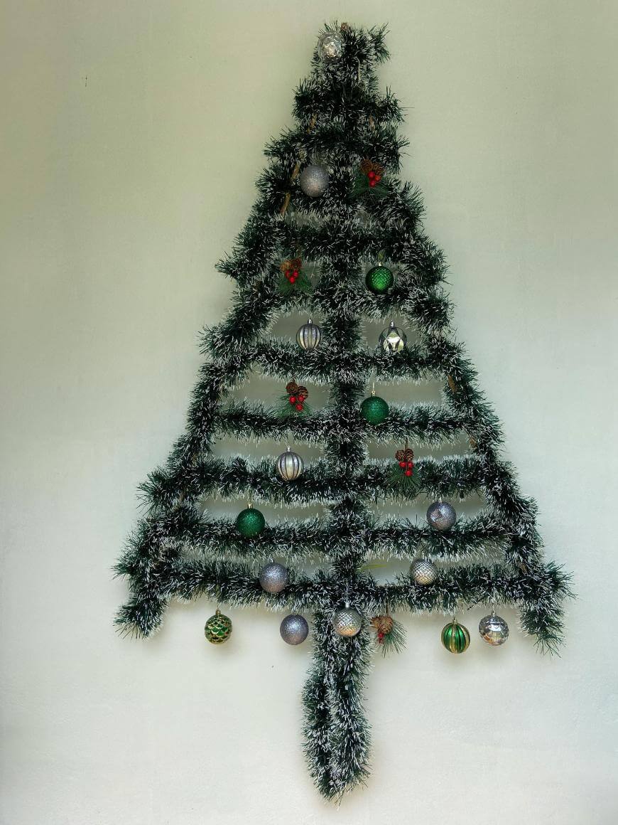 A Christmas tree shape made from green tinsel is arranged on a wall, decorated with green, silver, and red ornaments, resembling a festive wall display.