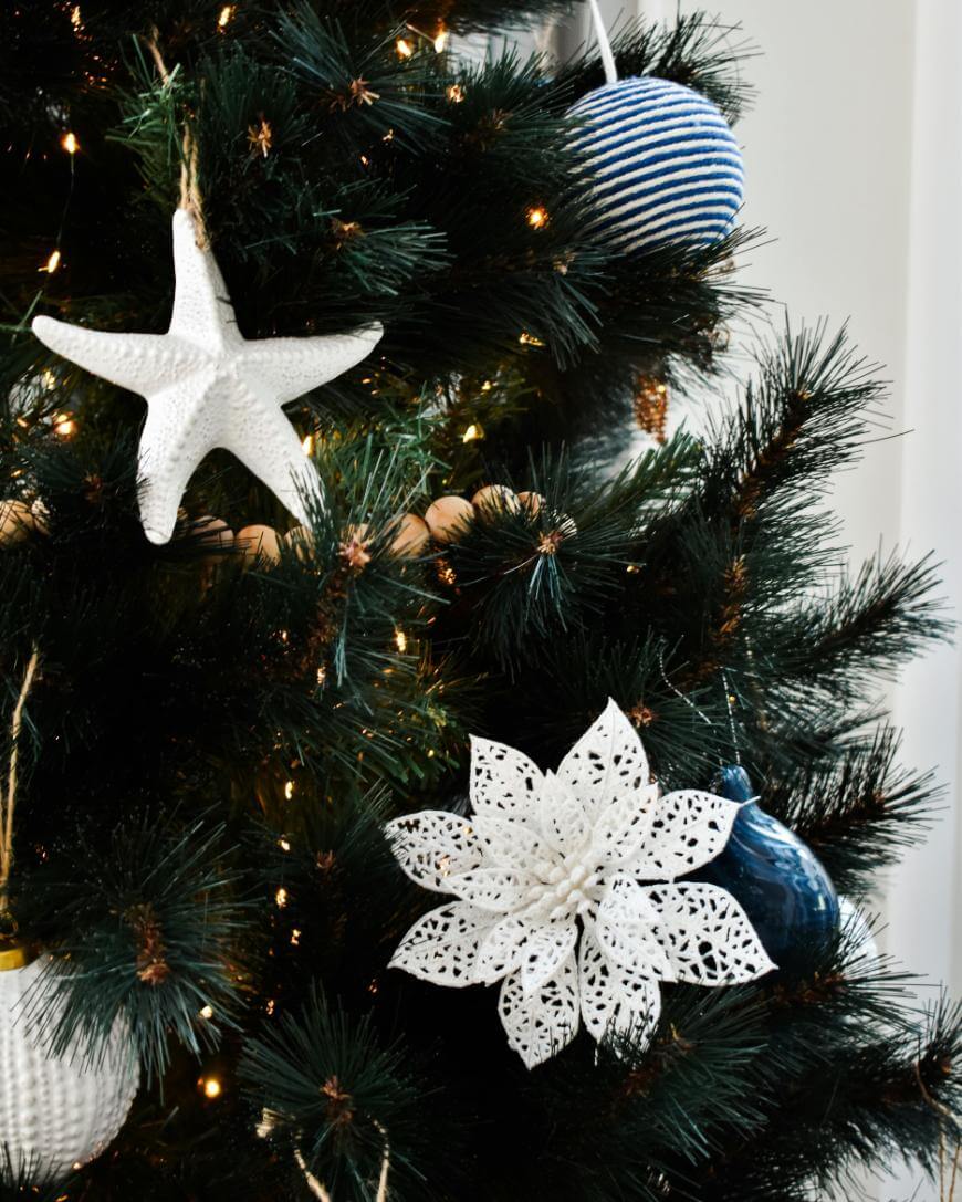 A close-up of a Christmas tree decorated with white starfish, a large white poinsettia ornament, and blue and white striped and solid blue round ornaments among green pine needles and string lights.