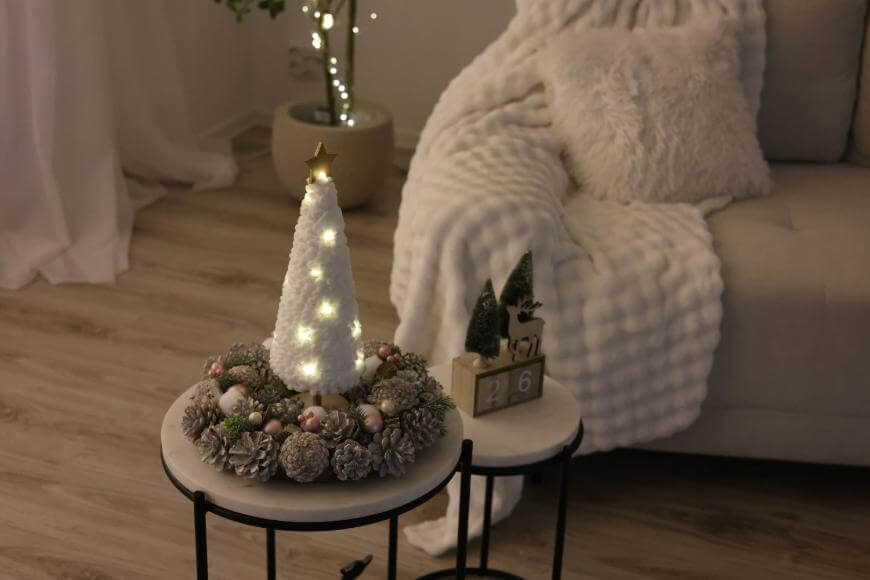 A cozy living room with a white textured blanket on a sofa. A round table displays a white cone Christmas tree with lights, surrounded by pinecones and ornaments. A small wooden calendar shows December 26.