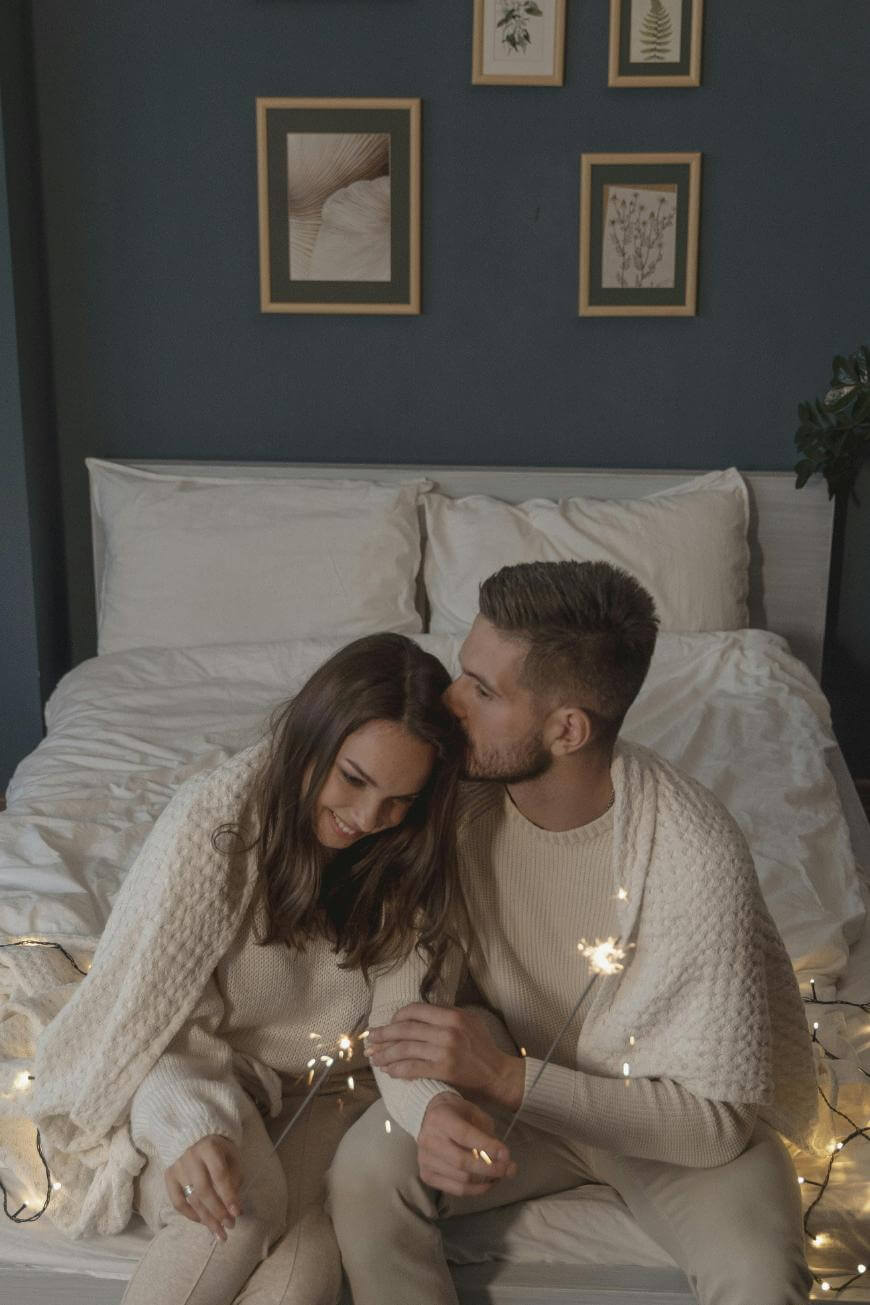 A couple sits on the edge of a bed wrapped in cozy sweaters, holding sparklers and smiling. The bed is neatly made with white linens, and framed artwork hangs on the dark blue wall behind them.