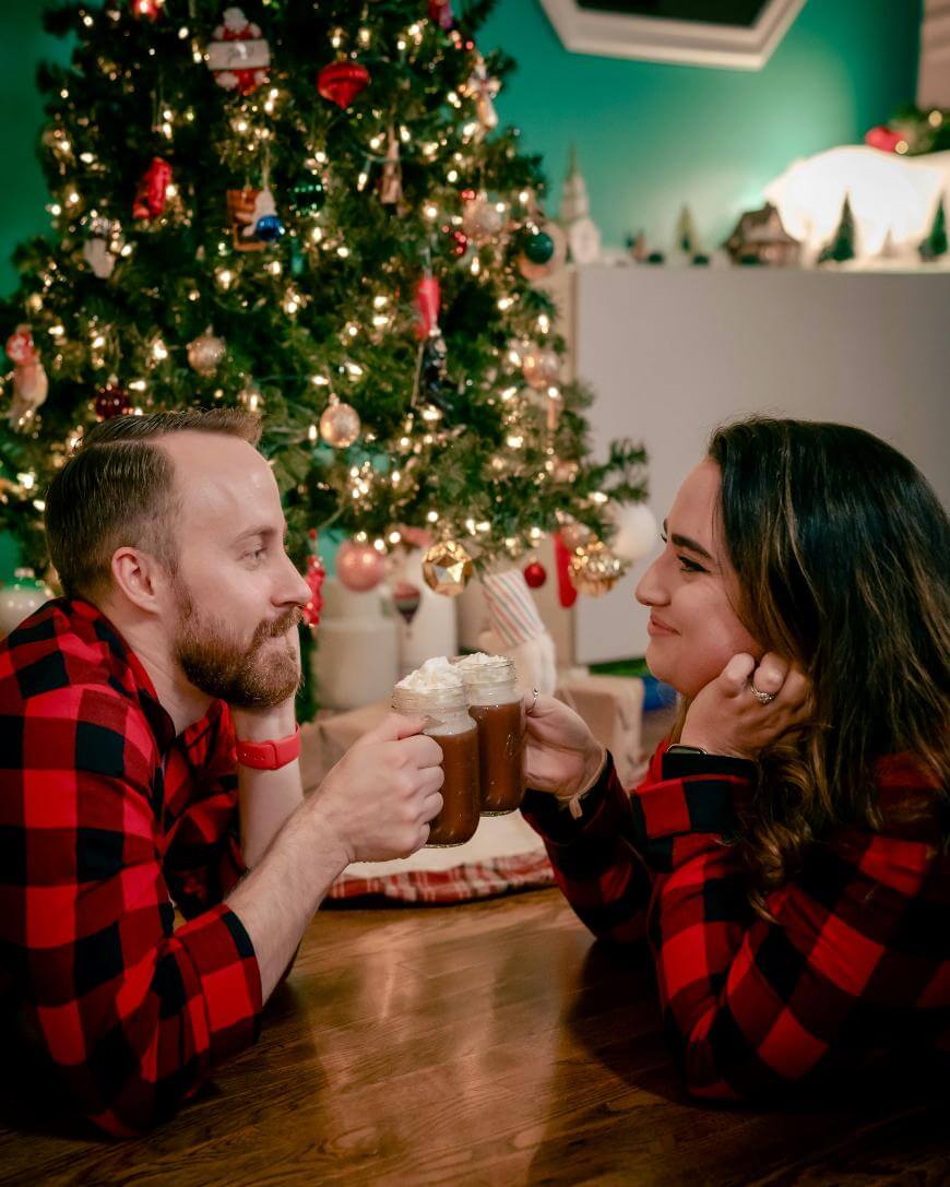 A man and woman in matching red and black plaid pajamas toast mugs of hot chocolate with whipped cream, smiling at each other in front of a decorated Christmas tree.