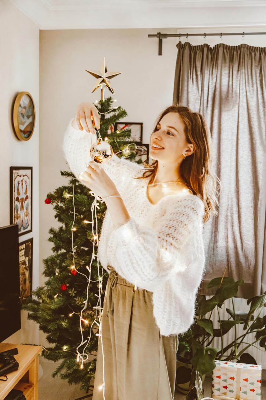 A woman in a white sweater decorates a Christmas tree with ornaments and string lights in a cozy living room. Wrapped gifts sit on the floor, and sunlight streams in through the window.