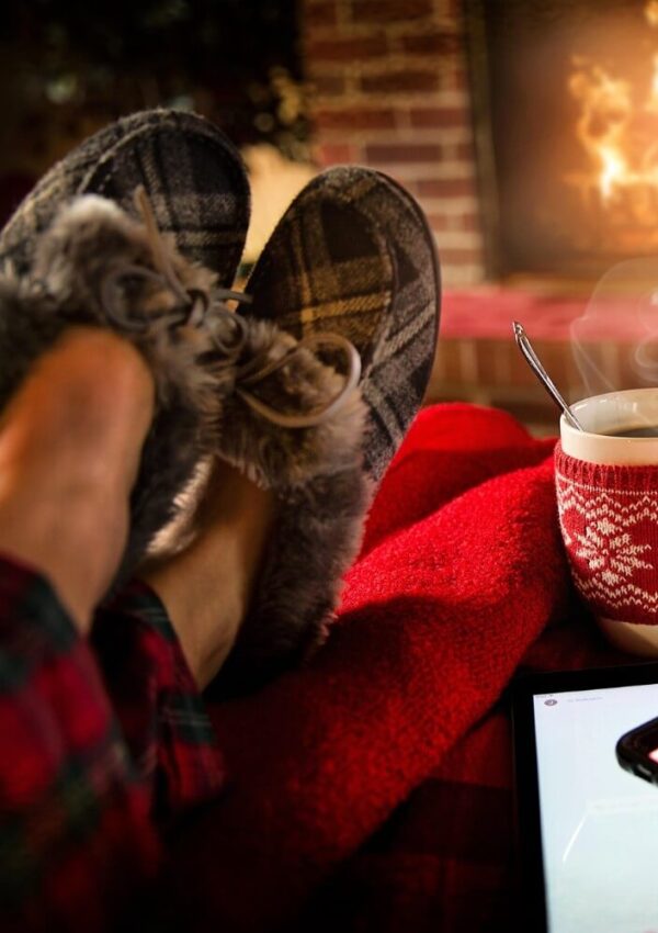A person wearing plaid slippers and pajama pants rests their feet on a red blanket near a fireplace. A mug with a knitted cozy and a spoon sits nearby, with steam rising from the drink.