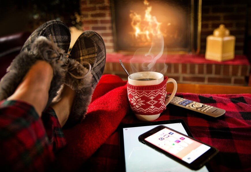 A person wearing plaid pajamas and slippers relaxes by a cozy fireplace with a steaming mug, a tablet, and a TV remote on a red blanket.